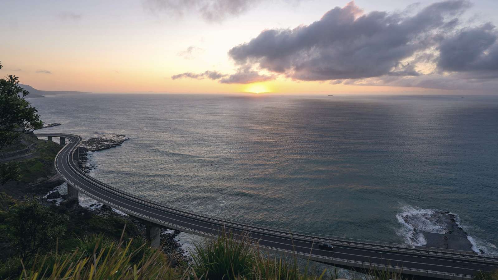 The Sea Cliff Bridge curves out over the sea below forested cliffs, with the sun low behind clouds on the horizon.