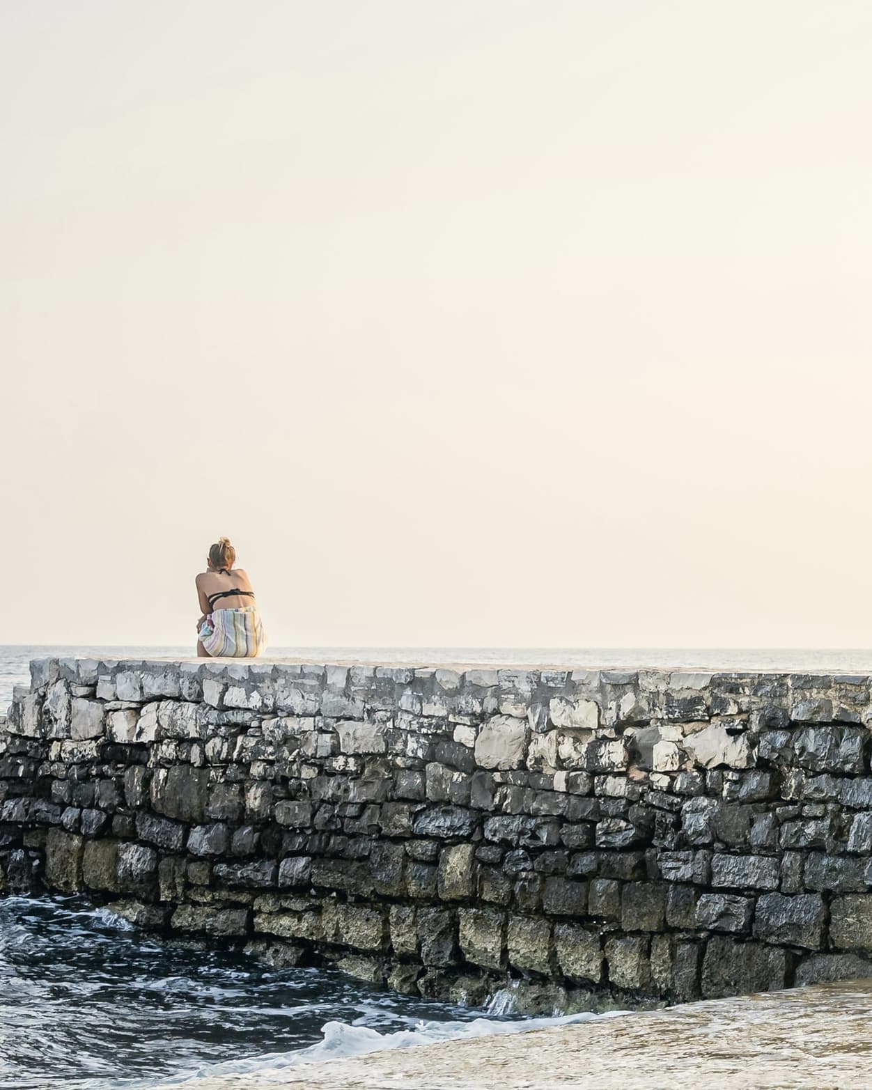 Stone steps lead down from an old breakwater into a calm sea under a pale sky, with one person sitting at the far end.
