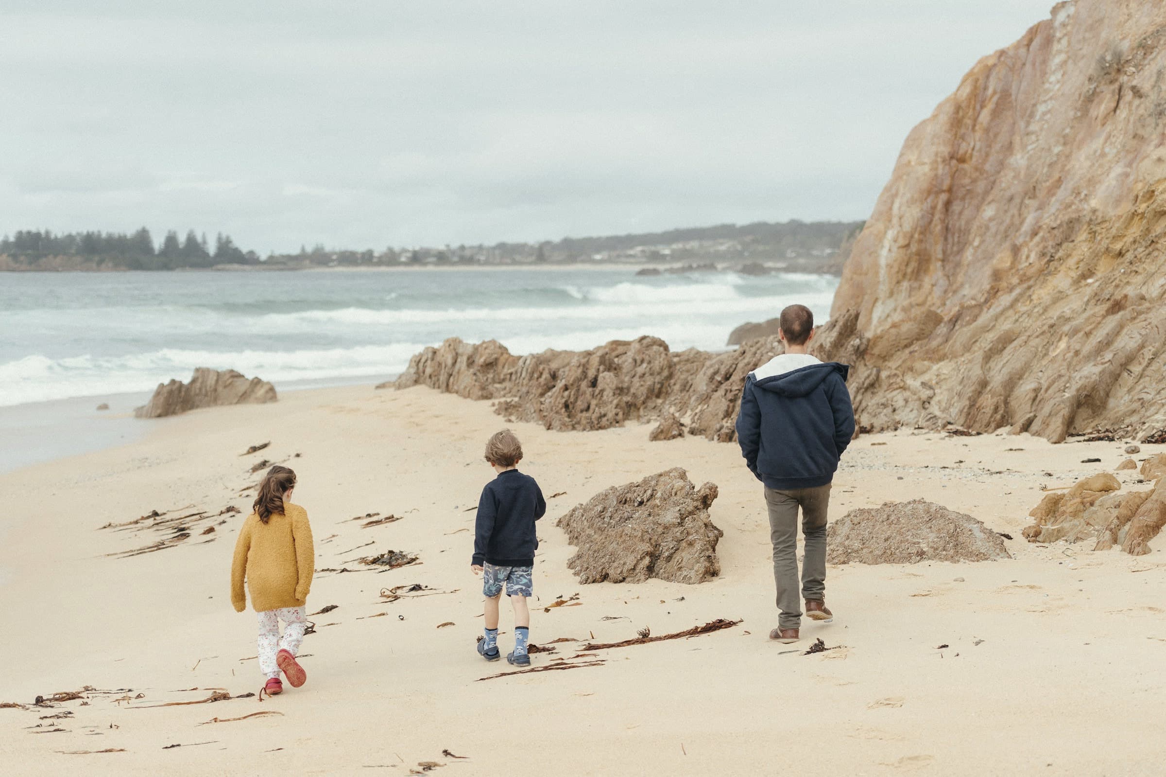 An adult walks along a sandy beach with two young children, past sandstone rocks and the surf.